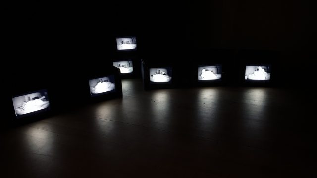 Installation detail: 7 small monitors on the floor in a dark room with images of McLeod sleeping