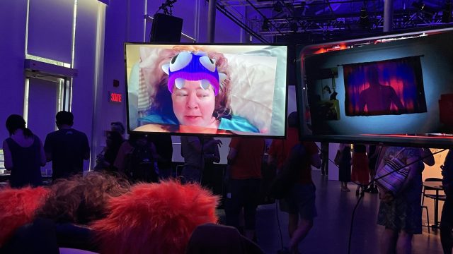 A video installation from Dayna McLeod's In Dreams that shows 2 monitors, one with Dayna's head wearing a muppet like hat, and the other with a sinister shadow in a dark window.