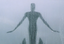 A sculptural figure with its arms outstretched as though ready to fly is behind a window covered in rain.