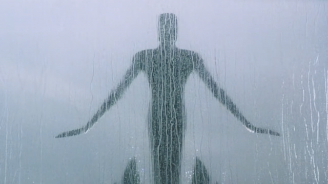 A sculptural figure with its arms outstretched as though ready to fly is behind a window covered in rain.