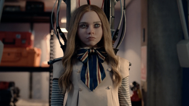 A life-sized doll with long blonde hair, a beige dress, and a blue, red, and white scarf tied in a bow, from the film M3GAN